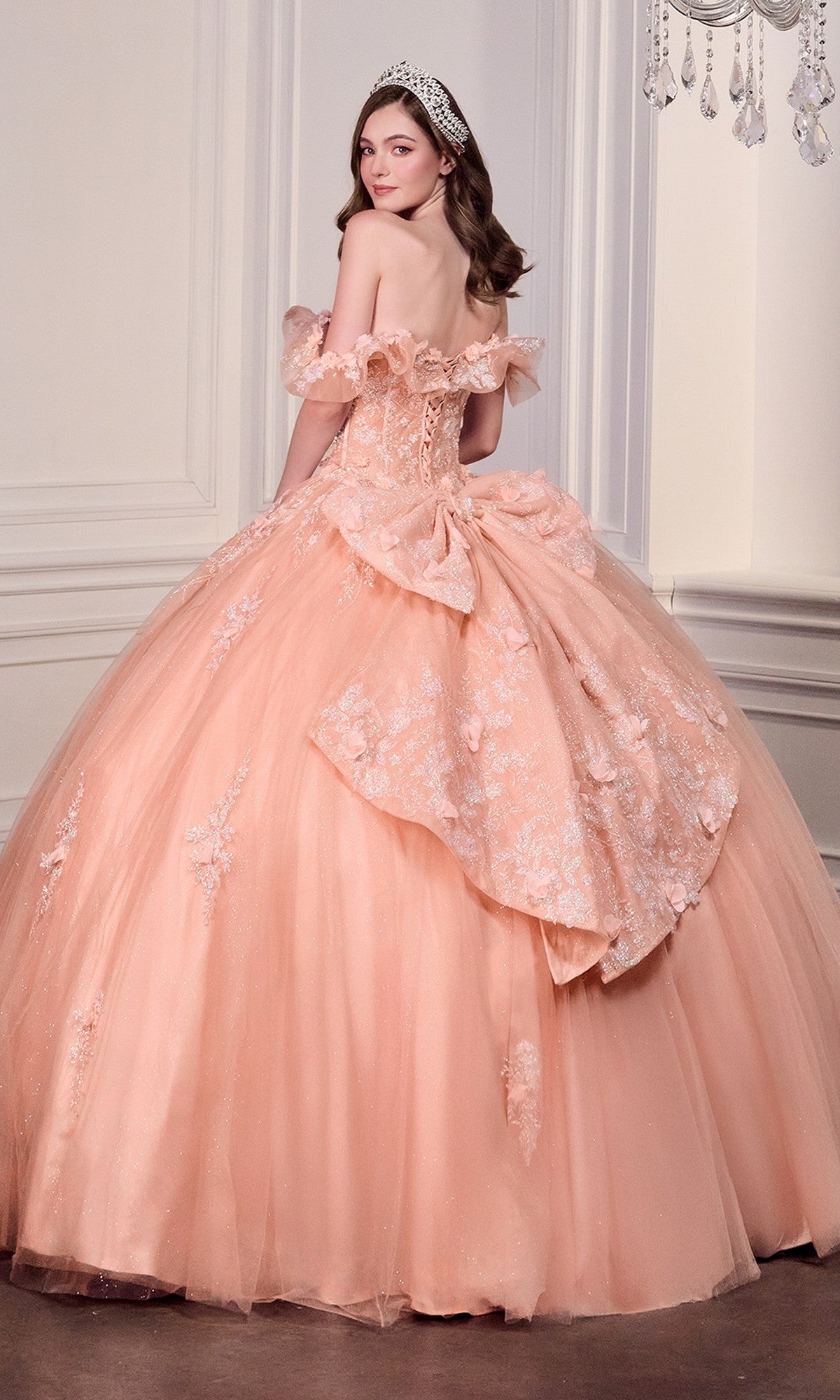 A young woman stands in an elegant room wearing the Quinceanera Dress 15731 By Ladivine, a peach off-the-shoulder gown with lace and 3D flowers, her tiara sparkling as she looks over her shoulder.